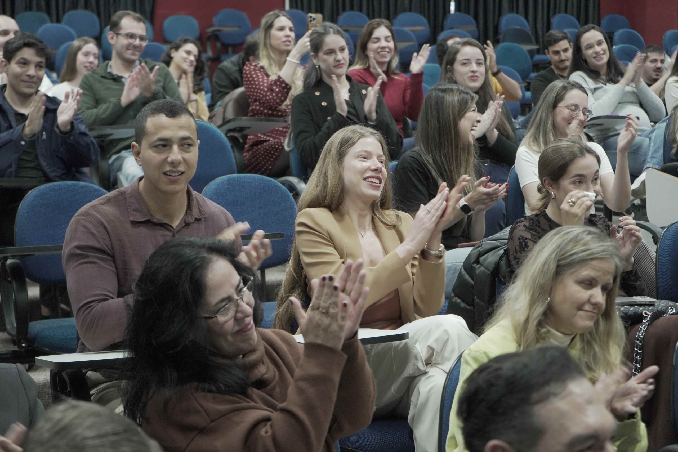 Auditório com estudantes, professores e convidados sentados em cadeiras azuis. Os participantes sorriem e aplaudem durante a Aula da Saudade da primeira turma de Medicina da UniFAJ, em um momento de celebração e reconhecimento pela trajetória acadêmica.