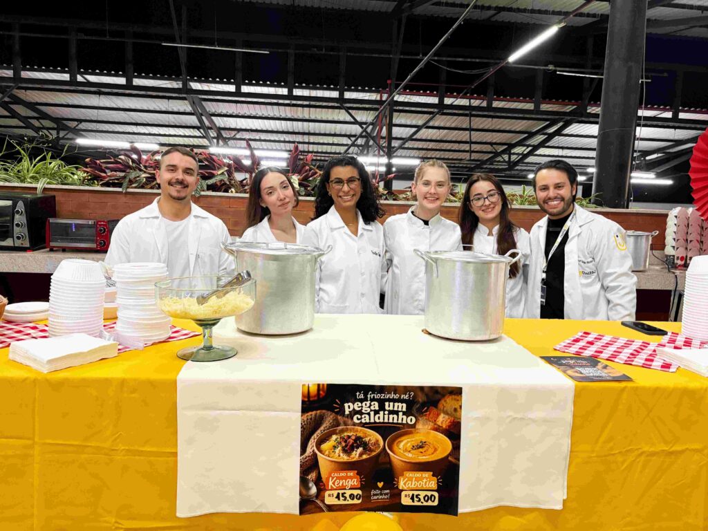 Estudantes de Nutrição, usando jalecos brancos, posam sorrindo atrás de um estande de gastronomia. Sobre a mesa há panelas, tigelas, pratos descartáveis e um cartaz divulgando caldos de kenga e de cabotiã. O ambiente é um festival gastronômico realizado à noite.