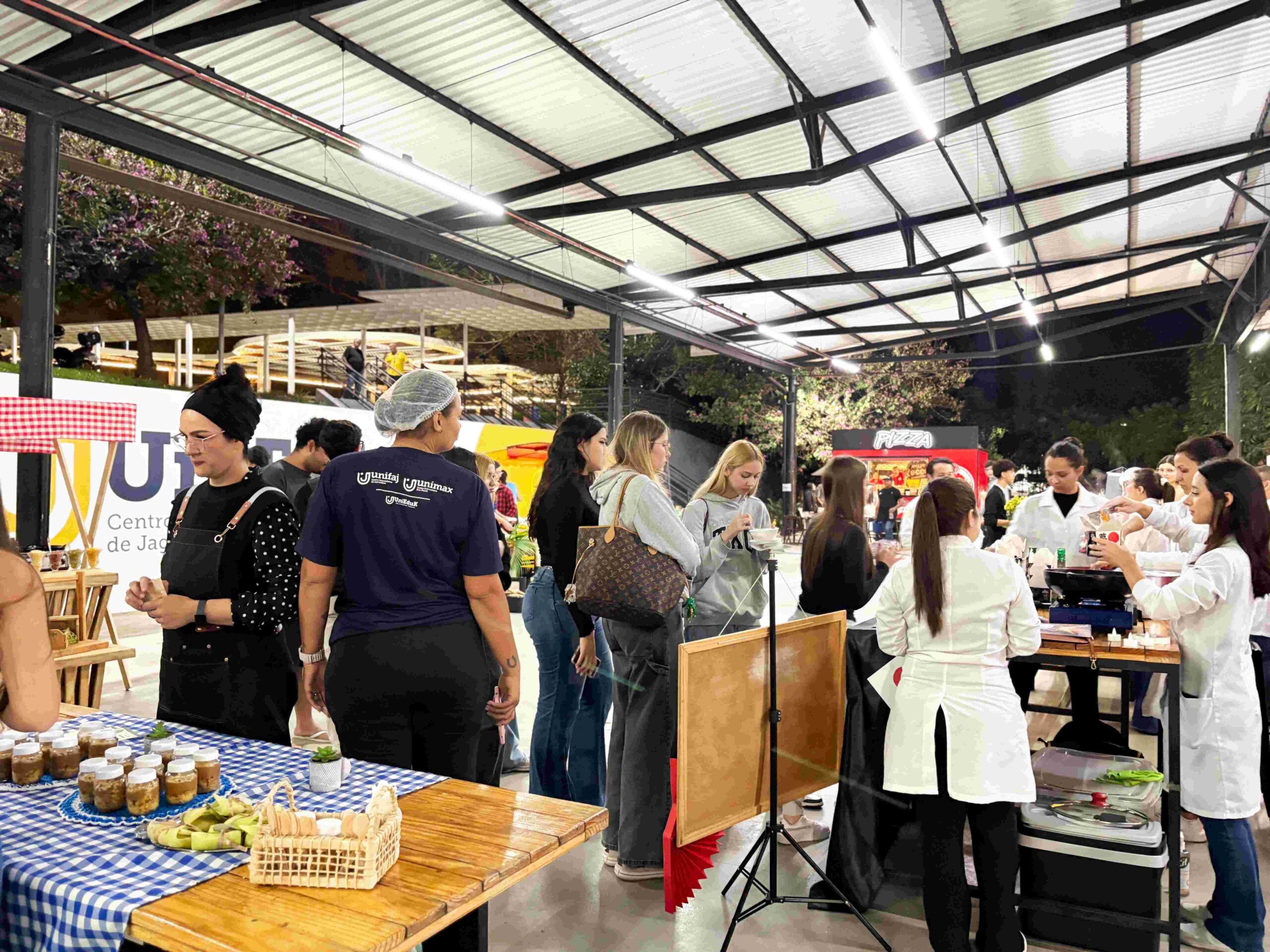 Visitantes circulam por diferentes estandes de gastronomia em um espaço coberto. Estudantes de jaleco servem preparações e apresentam os pratos ao público, enquanto mesas com alimentos e decorações temáticas compõem o ambiente do festival.